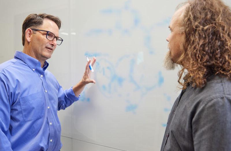 博彩平台网址大全 scientists Brett Crawford and Dan Wendt talking by a whiteboard with a scientific formula